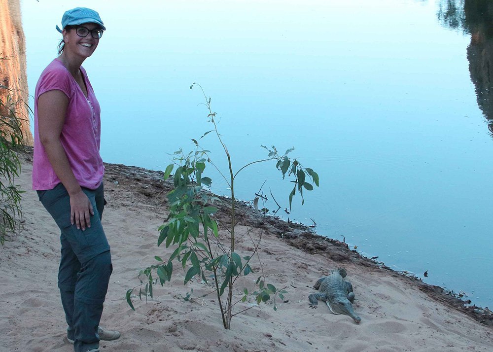 A woman in a pink shirt and blue hat stands near a body of water with a sweet water crocodile next to her on the sand.