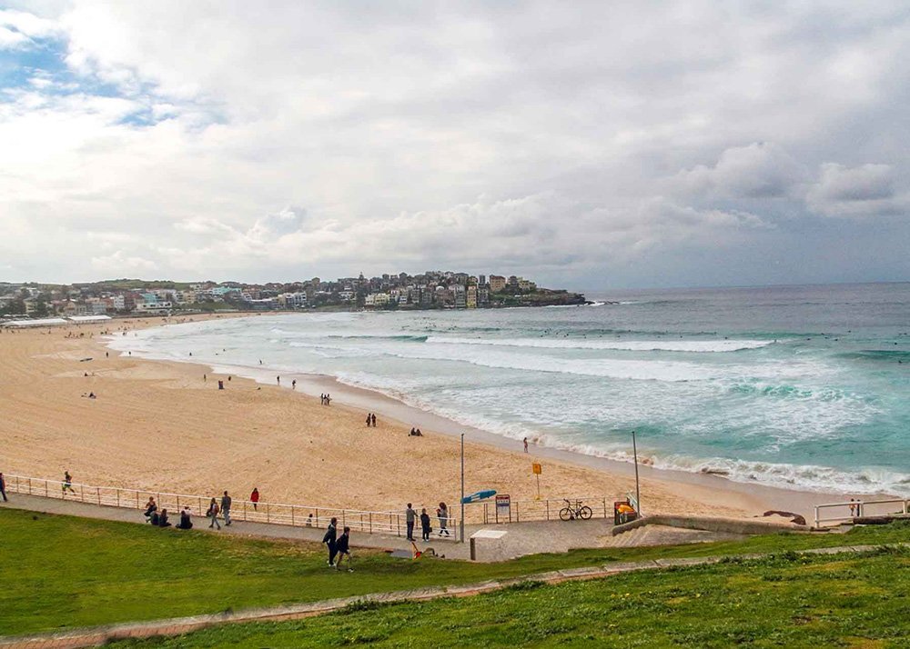 Wide view of Bondi Beach with gentle waves crashing onto the sandy shore, a few people walking and relaxing, and a distant view of houses along the coastline under a mostly cloudy sky.
