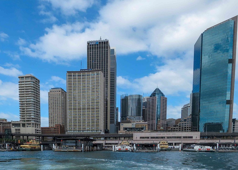 View of modern skyscrapers at Circular Quay in Sydney, with high-rise buildings including the AMP tower, seen from across the water under a blue sky with some clouds.