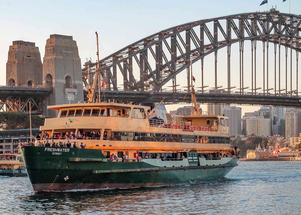 Sydney ferry named 'Freshwater' cruising on the water with the Sydney Harbour Bridge in the background, carrying passengers and framed by the city's skyline at dusk."