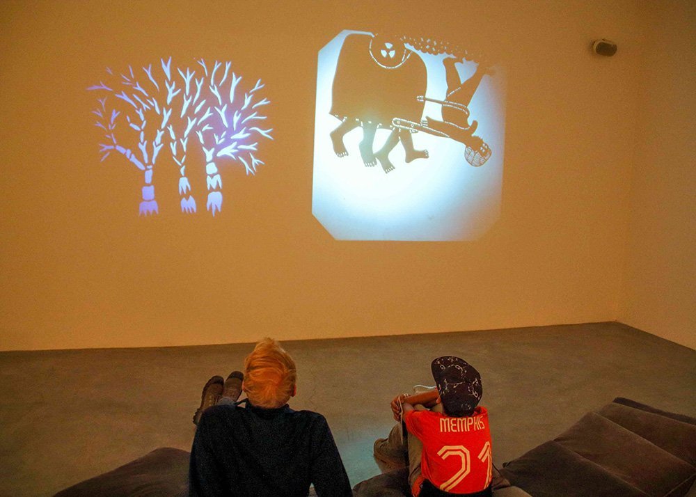 A father and child sitting on a couch watching shadow puppet projections on a museum wall, featuring a tree on the left and a whimsical figure on the right