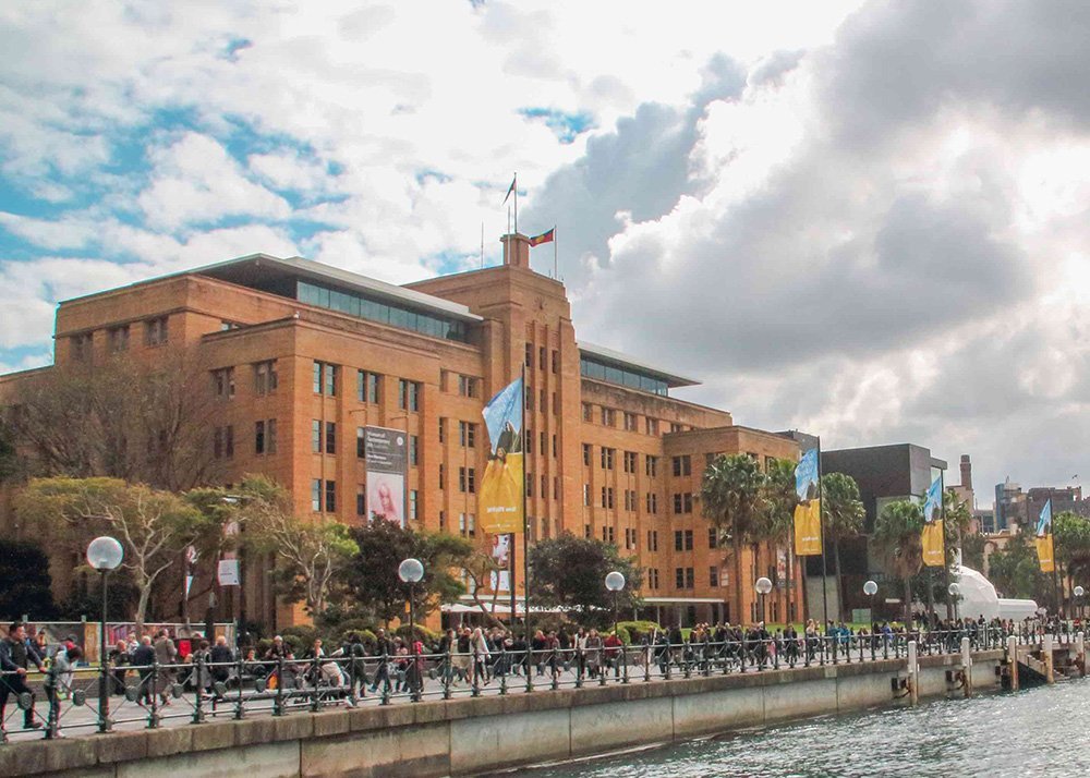 Sydney Museum of Contemporary Art, a large brick building with banners on its facade, a busy pedestrian walkway in front, and a partly cloudy sky.