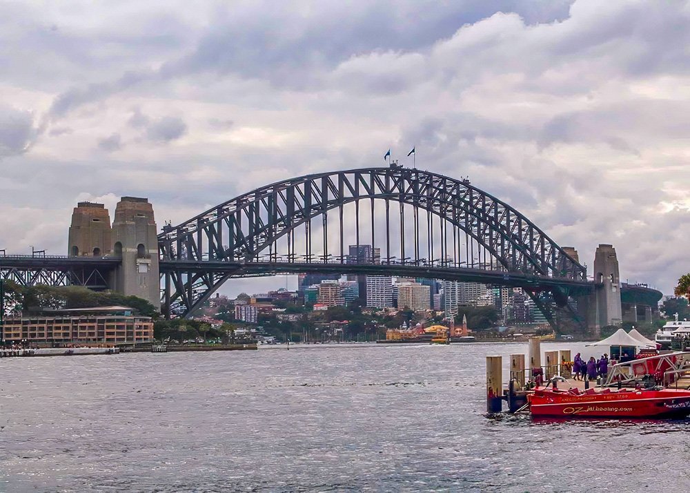 Sydney Harbour Bridge spanning across the water with city buildings in the background and a cloudy sky overhead.