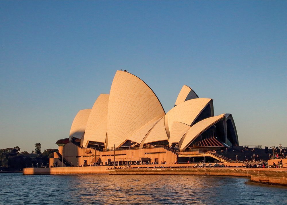 Sydney Opera House illuminated by the golden light of the setting sun, its iconic sails standing out against a clear blue sky.