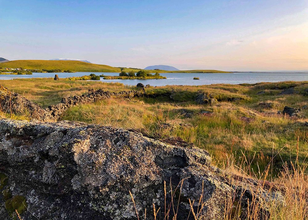 Expansive view of a grassy landscape with rocky outcrops, Lake Myvatn in the distance, and rolling hills under a clear sky at sunset.
