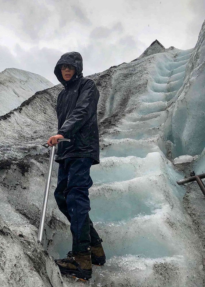 A teenage boy in a black rain jacket and boots, holding an ice pick while hiking up a staircase carved into a glacier under an overcast sky.