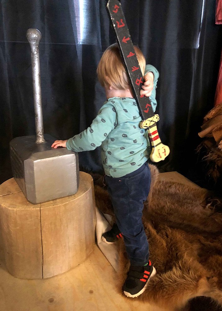 Young child holding a toy Viking sword standing next to a large Thor hammer prop inside Valhalla restaurant and saga center