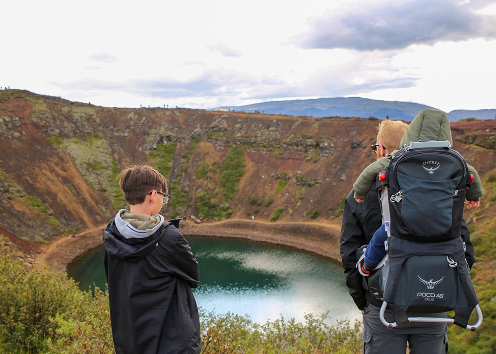 A man carrying a baby in a back carrier and a teen standing next to them are staring into the lake in the Kerid Crater in Iceland
