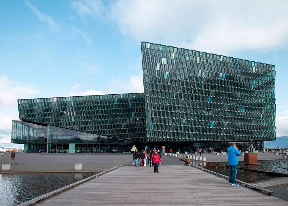 Harpa music hall is a contemporary glass building with a unique geometric design, located by the water with people walking on a wooden boardwalk.
