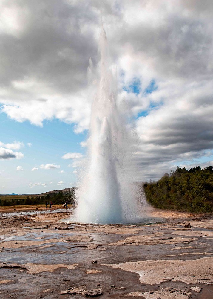 Strokkur geyser in Iceland erupting with a powerful jet of water into the sky, surrounded by a rocky landscape and a few spectators.