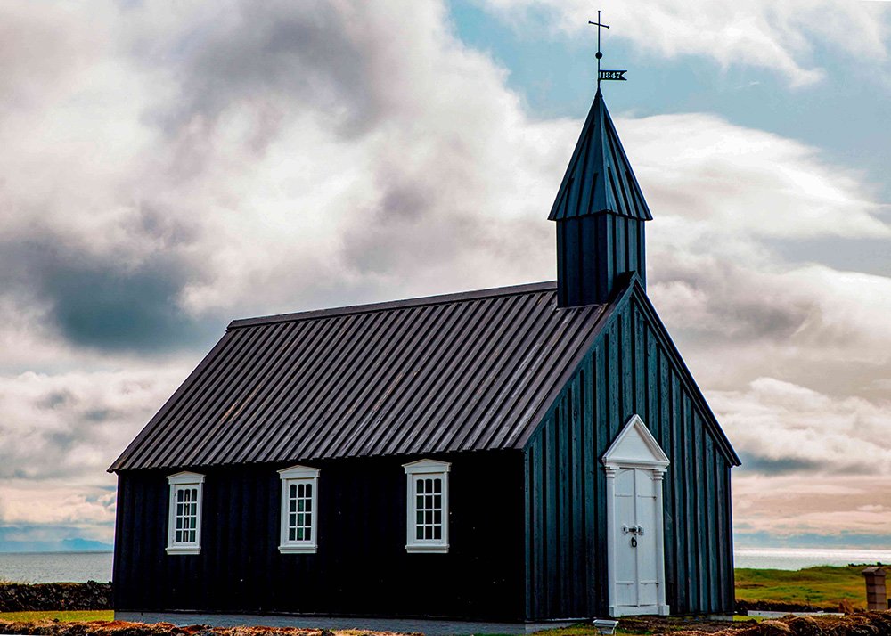 Black wooden church with a white door and windows, situated on a grassy plain under a cloudy sky