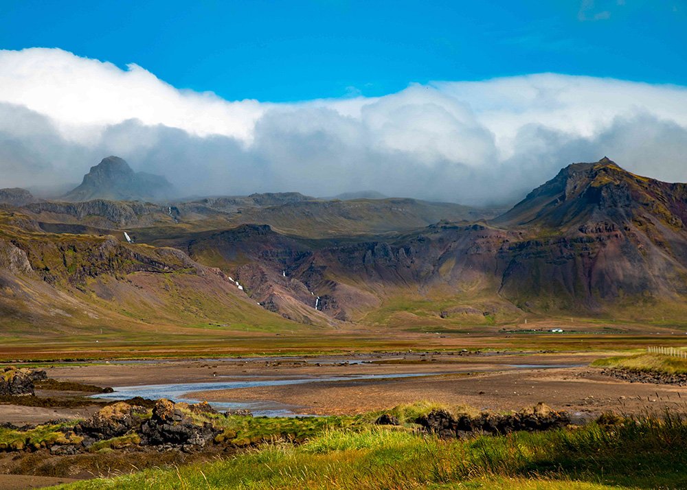 A scenic view of mountains with low-hanging clouds partially covering the peaks, grassy fields in the foreground, and small waterfalls cascading down the slopes.