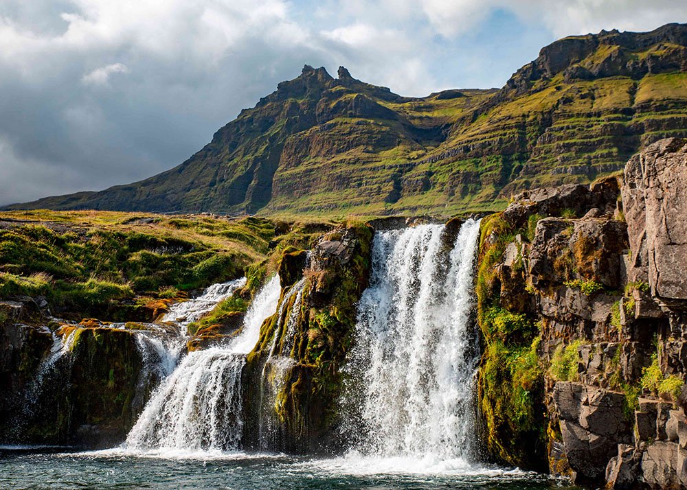 A picturesque waterfall cascading over moss-covered rocks, with a mountainous backdrop under a partly cloudy sky.