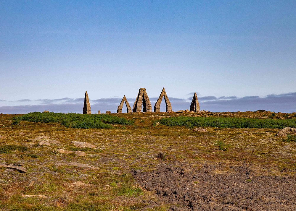 The stone arch structures known as the Arctic Henge stand alone in a grassy, rocky landscape under a clear blue sky.