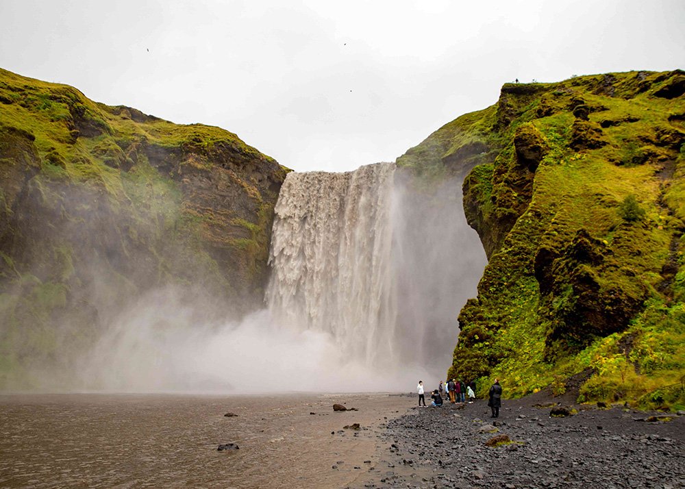 A group of people standing near the base of the powerful Skogafoss waterfall surrounded by lush green cliffs.