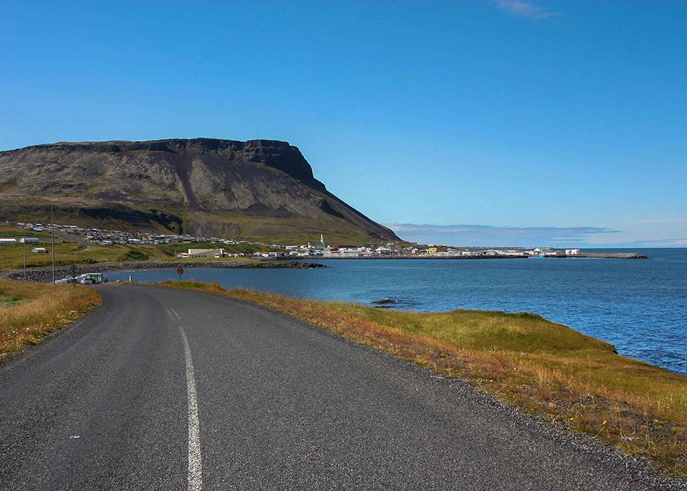 A scenic coastal road in Olafsvik, Iceland, with the town and mountains in the distance.