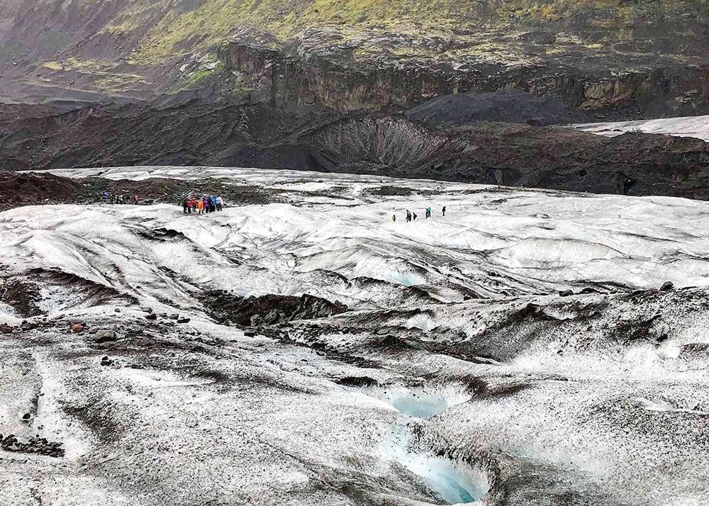 A group of hikers walk across the icy expanse of Skaftafell glacier in Iceland, surrounded by rugged, snow-covered terrain. The hikers are bundled up in warm clothing, and the glacier stretches out towards rocky cliffs.