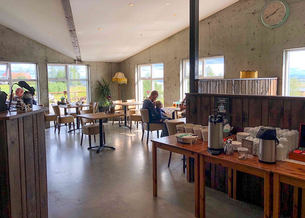 The interior of Efstidalur Farm restaurant with wooden tables, chairs, and large windows letting in natural light. Customers are seated, enjoying their drinks and conversations in a warm and inviting atmosphere.