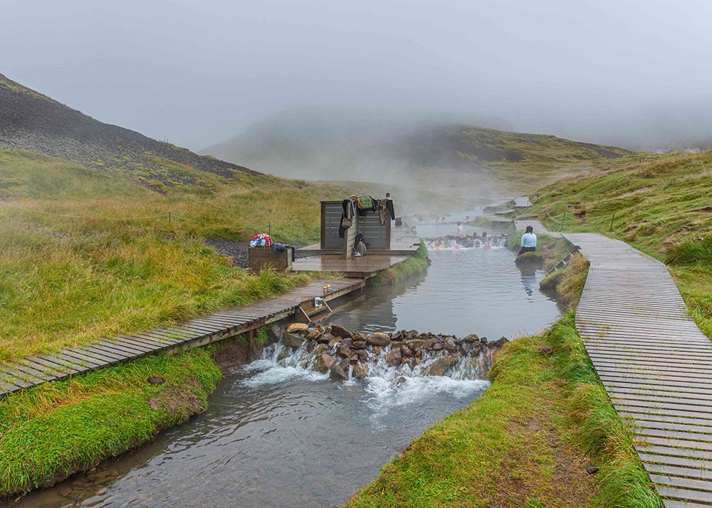 Scenic geothermal Reykjadalur in Iceland with steaming hot springs and rugged, moss-covered lava rocks. Wooden pathways lead visitors through the area, with a misty, ethereal atmosphere.