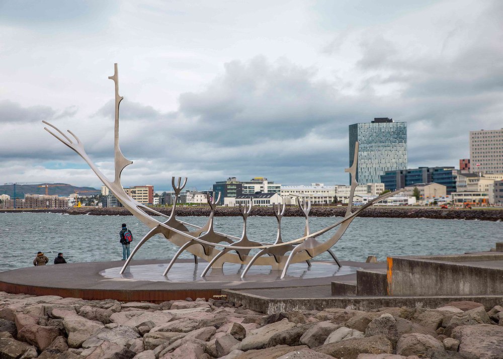 The Sun Voyager sculpture in Reykjavik, Iceland, representing a dreamboat or an ode to the sun. The sculpture is set against the backdrop of the city's skyline and the ocean, with cloudy skies above.