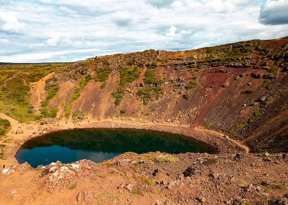 The Kerid volcanic crater in Iceland, with its steep, colorful walls and a blue-green lake at the bottom. The rocky, reddish slopes surround the serene water, creating a striking natural feature.