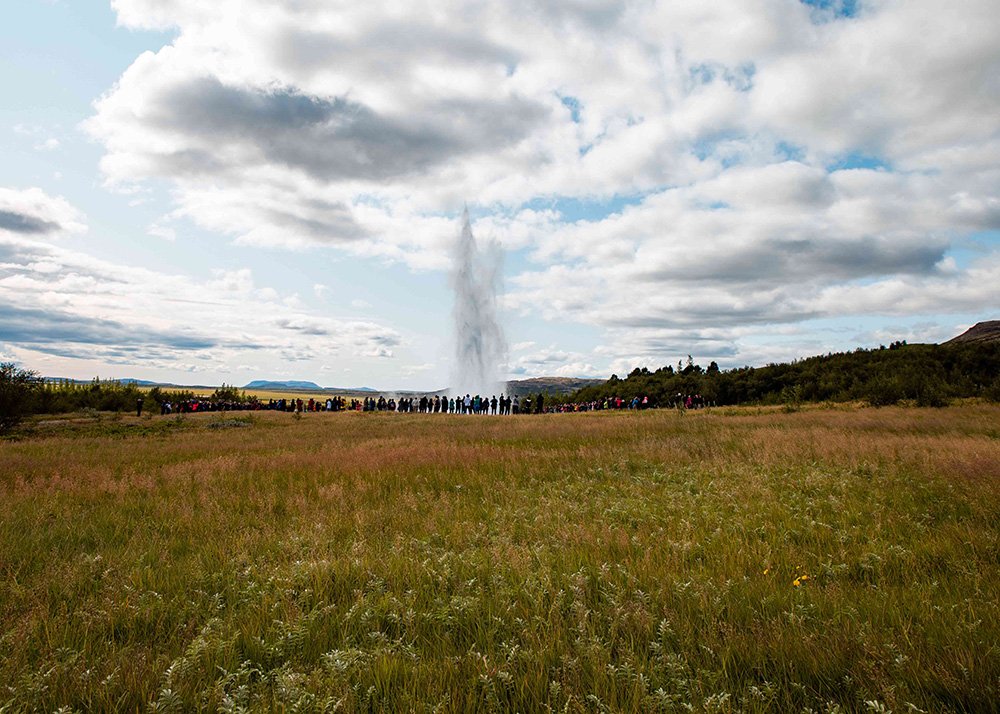 Strokkur geyser erupts in a field in Iceland, sending a tall plume of water and steam into the sky. Tourists stand in a line at a distance, watching the natural spectacle unfold.