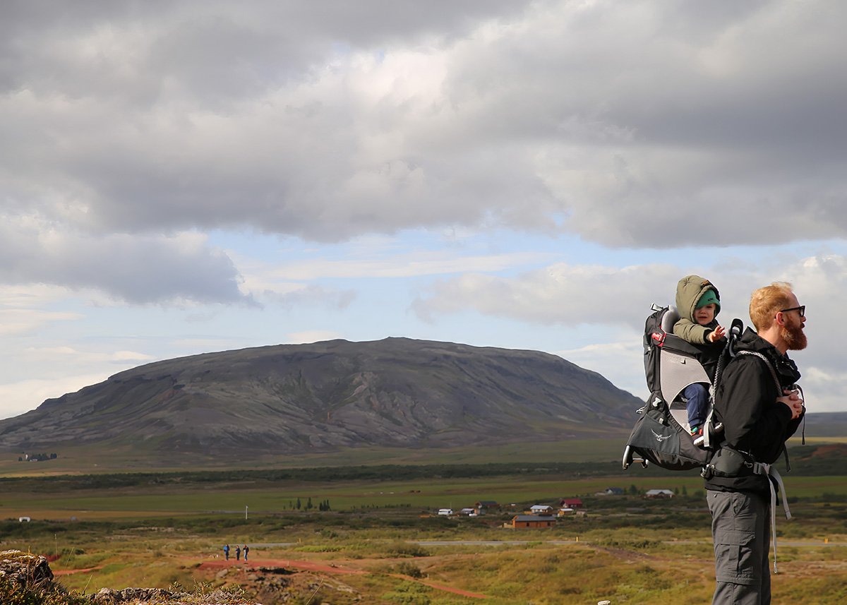 A father carries his baby in a back carrier. In the background Mount Búrfell can be seen. With a cloudy sky above signaling it's about to rain.
