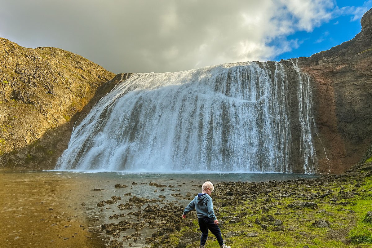A boy is walking on small rocks right in front of Thorufoss on a sunny day.
