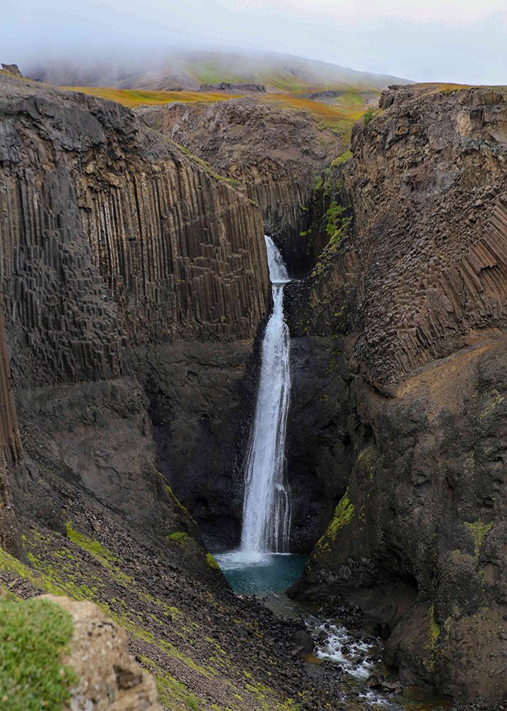 Litlanesfoss waterfall plunging down a cliff with basalt columns, surrounded by rugged terrain.
