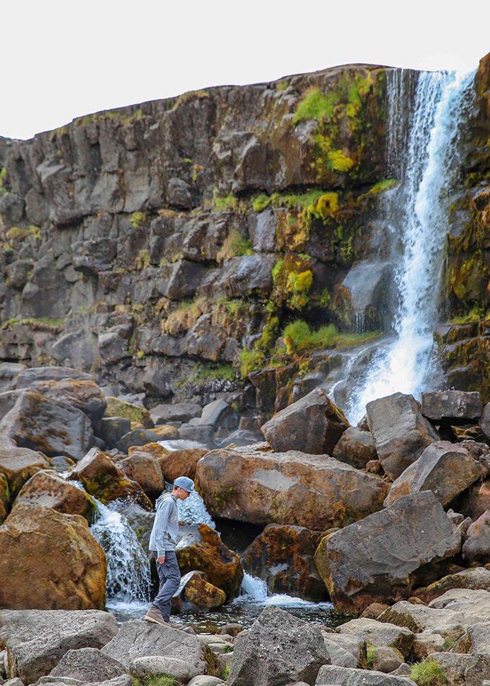 A person walking among large boulders near a waterfall cascading down a rocky cliff.