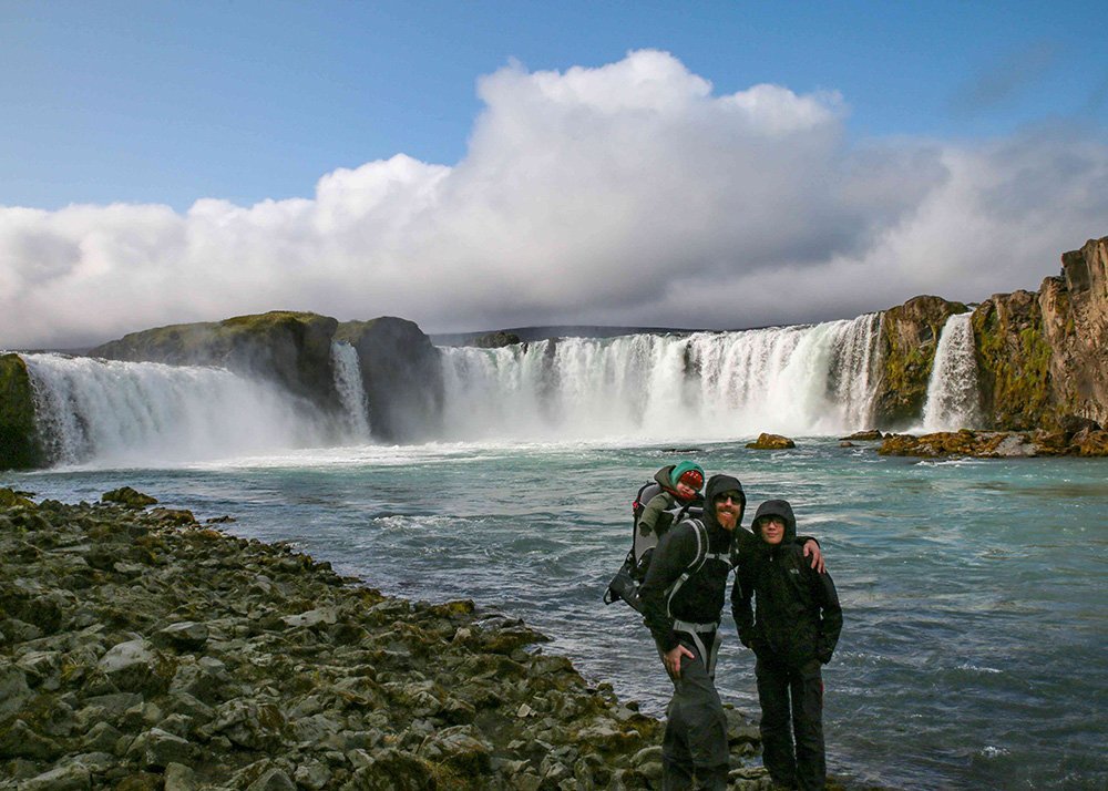 A family posing in front of the powerful Godafoss waterfall with mist rising from the water.