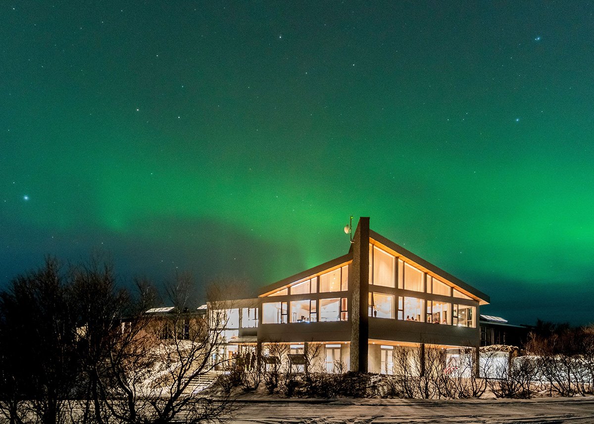 The contemporary glass-fronted building of Hotel Húsafell in West Iceland glows warmly under a dazzling green aurora borealis in a clear starry night sky, surrounded by bare winter trees and snowy ground.