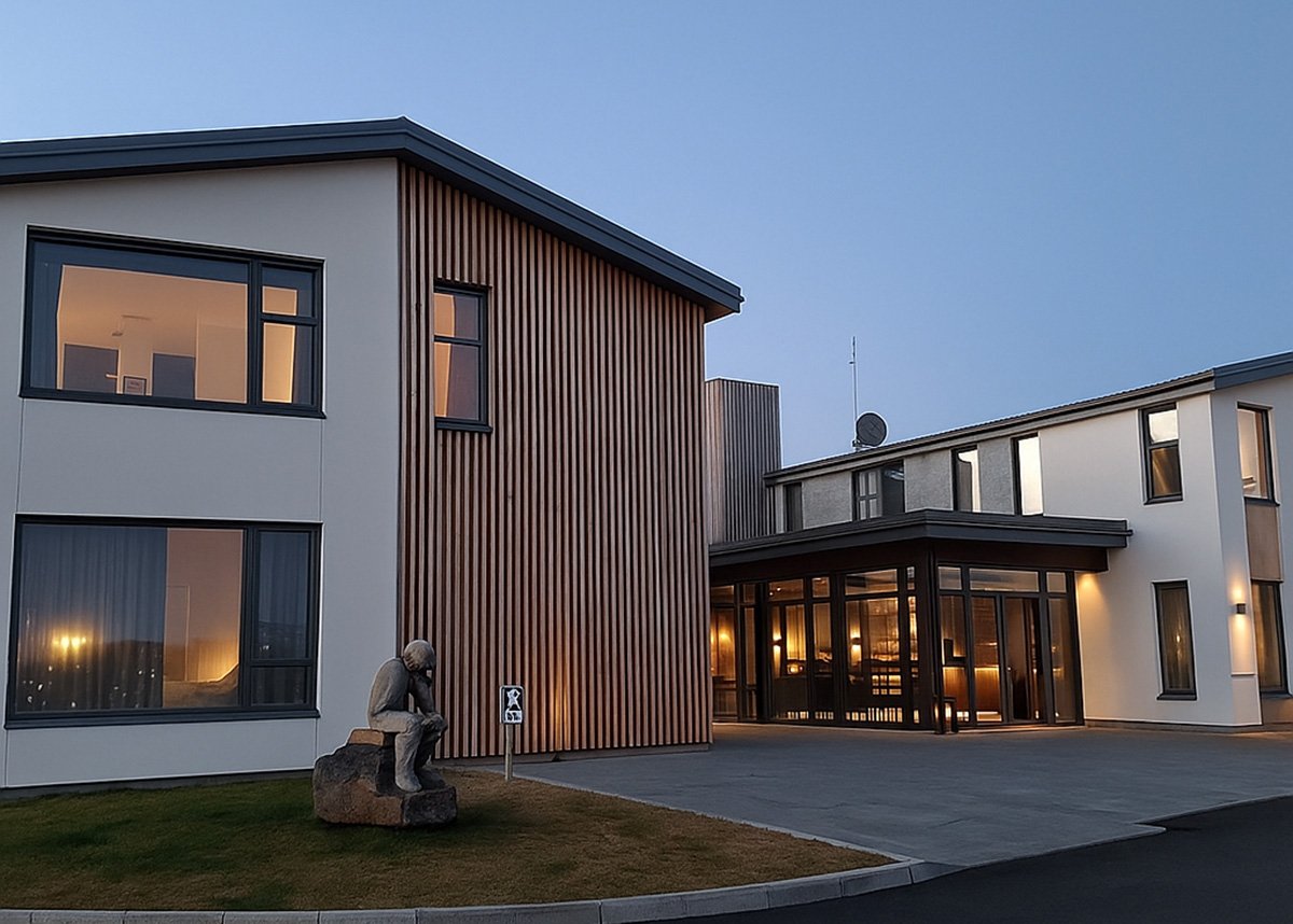 The sleek modern facade of Berjaya Mývatn Hotel with glowing interior lights, a wood-panel feature wall, and a contemplative statue on the lawn, photographed during dusk in North Iceland.