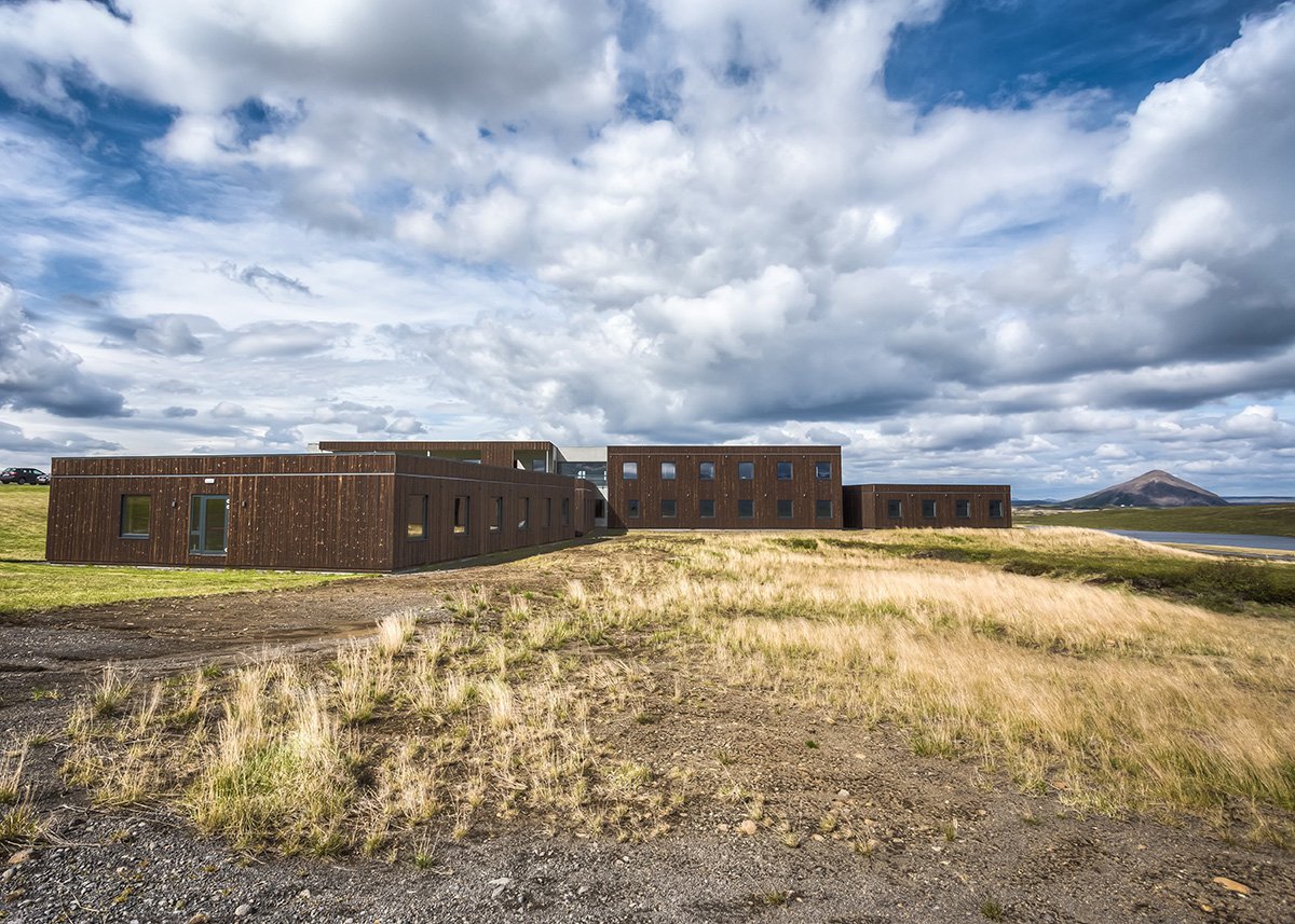 Hotel Laxá’s low, wooden buildings stretch across an open field near Lake Mývatn, North Iceland, under a strikingly cloudy sky with patches of blue, conveying a remote yet welcoming atmosphere.