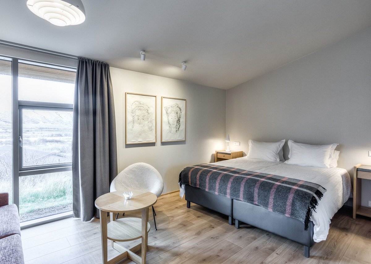 A stylish and serene room at Hotel Húsafell features minimalist decor, a large bed with crisp white linens, two delicate line-art portraits, and floor-to-ceiling windows offering views of the frosty Icelandic landscape.