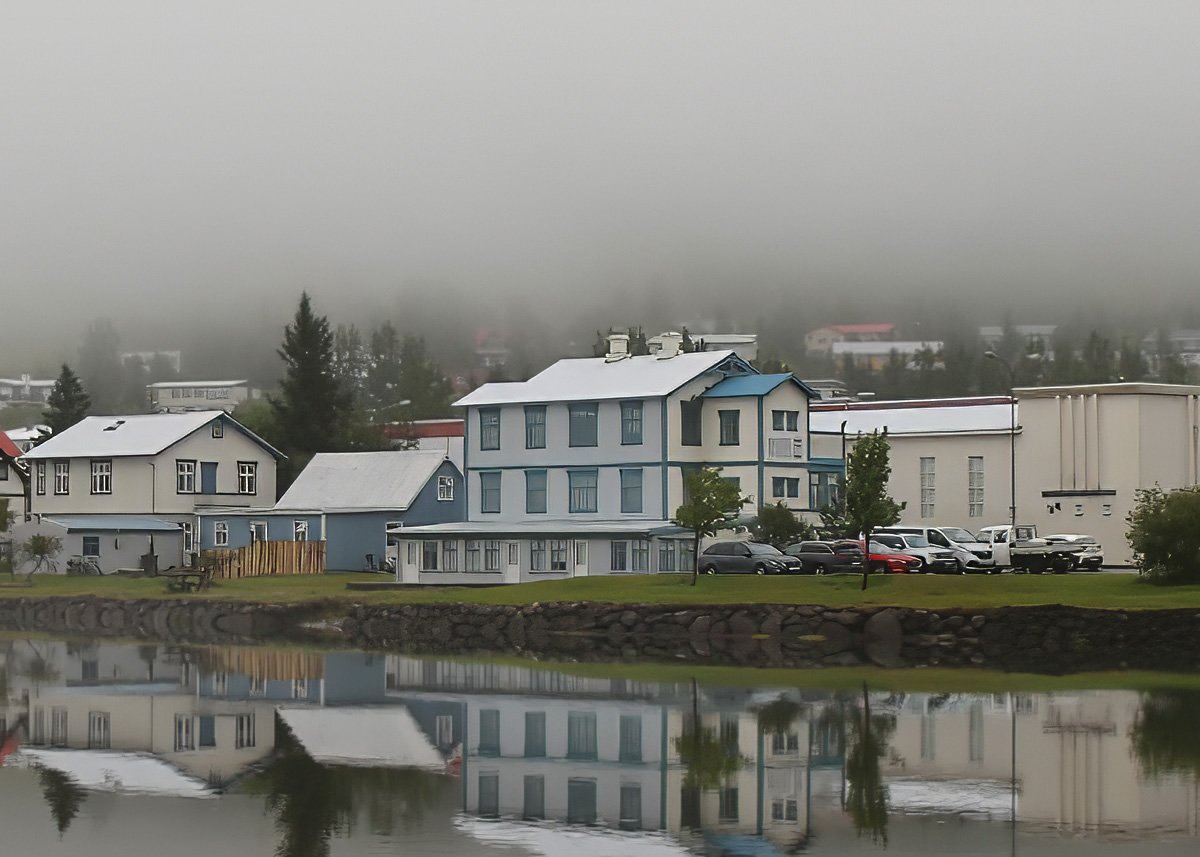 The blue-and-white exterior of Snæfell, Hotel by Aldan, reflected in the calm waters of a fjord in the East Iceland town of Seyðisfjörður, shrouded in light fog and backed by colorful houses and evergreen trees.