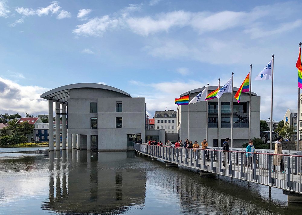 Reykjavik City Hall, a modern building on Tjörnin pond, with a bridge and rainbow flags displayed outside.