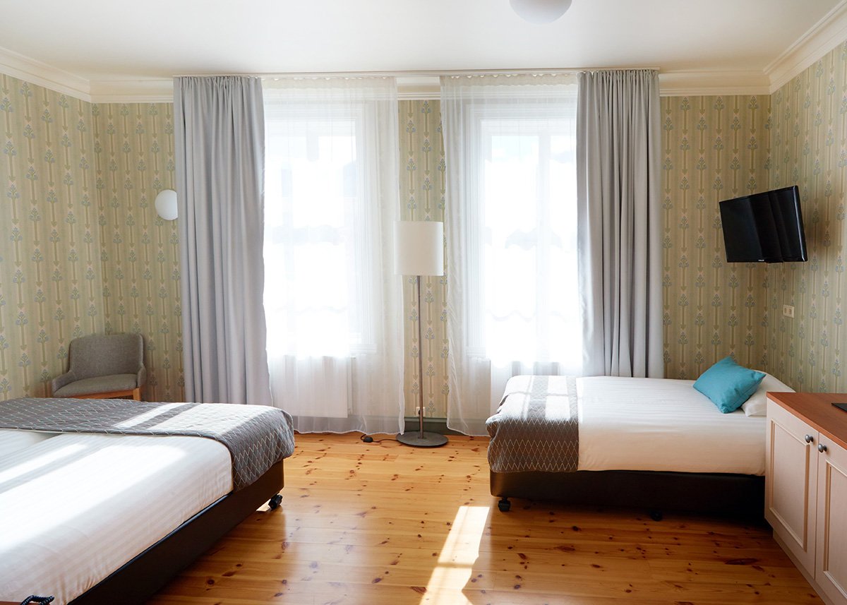 A sunlit, traditionally styled twin room at Fosshotel Eastfjords in East Iceland features a mix of classic wallpaper, natural wood floors, soft gray curtains, and modern bedding—perfect for families seeking comfort with a local touch.