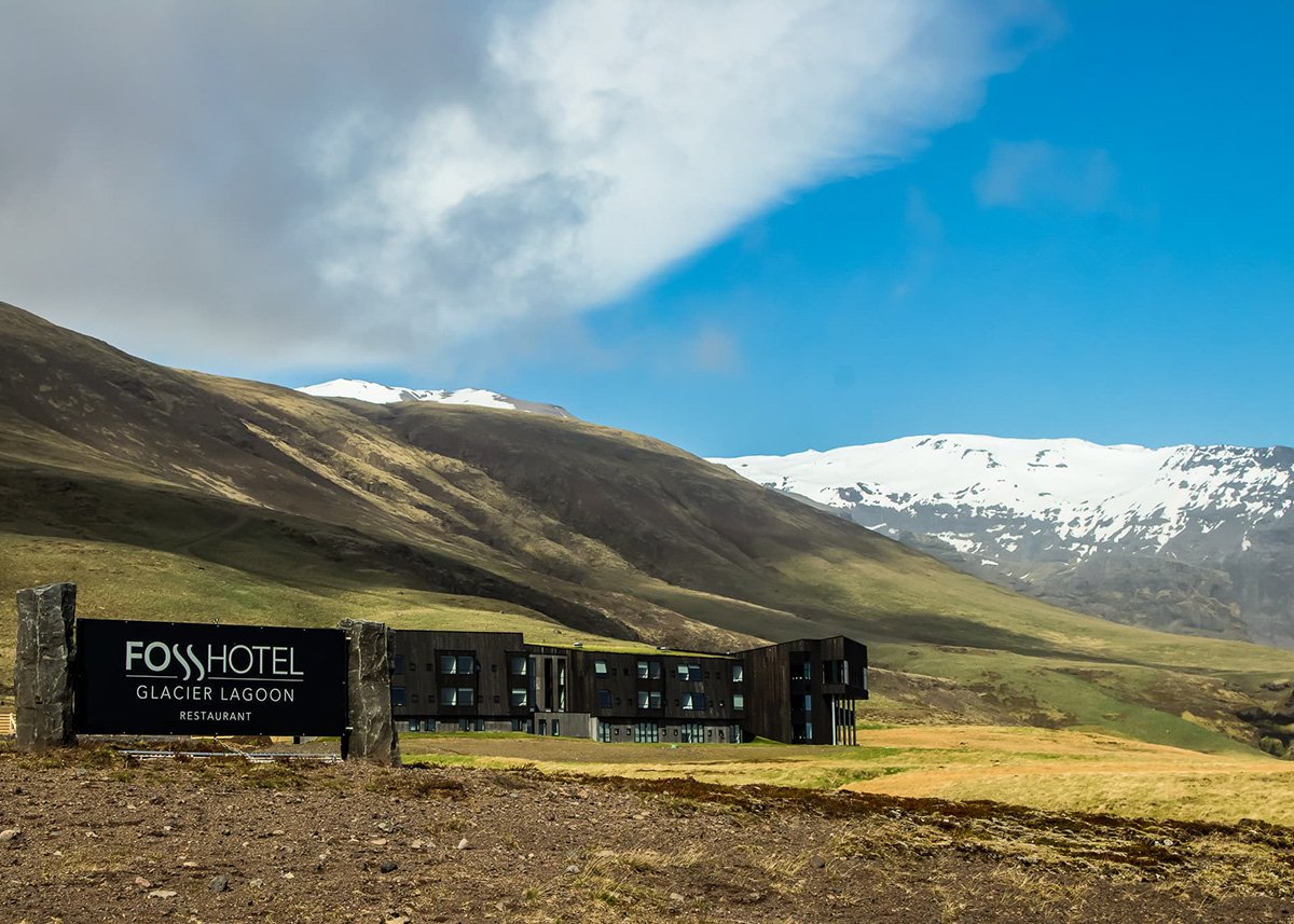 Fosshotel Glacier Lagoon near Skaftafell, South Iceland, stands boldly beneath rolling green hills and snow-covered peaks under a partially cloudy sky, with its dark, modern facade contrasting the wild, remote landscape.