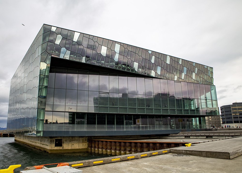 The modern glass Harpa concert hall with geometric patterns reflecting the cloudy sky and surrounding cityscape.