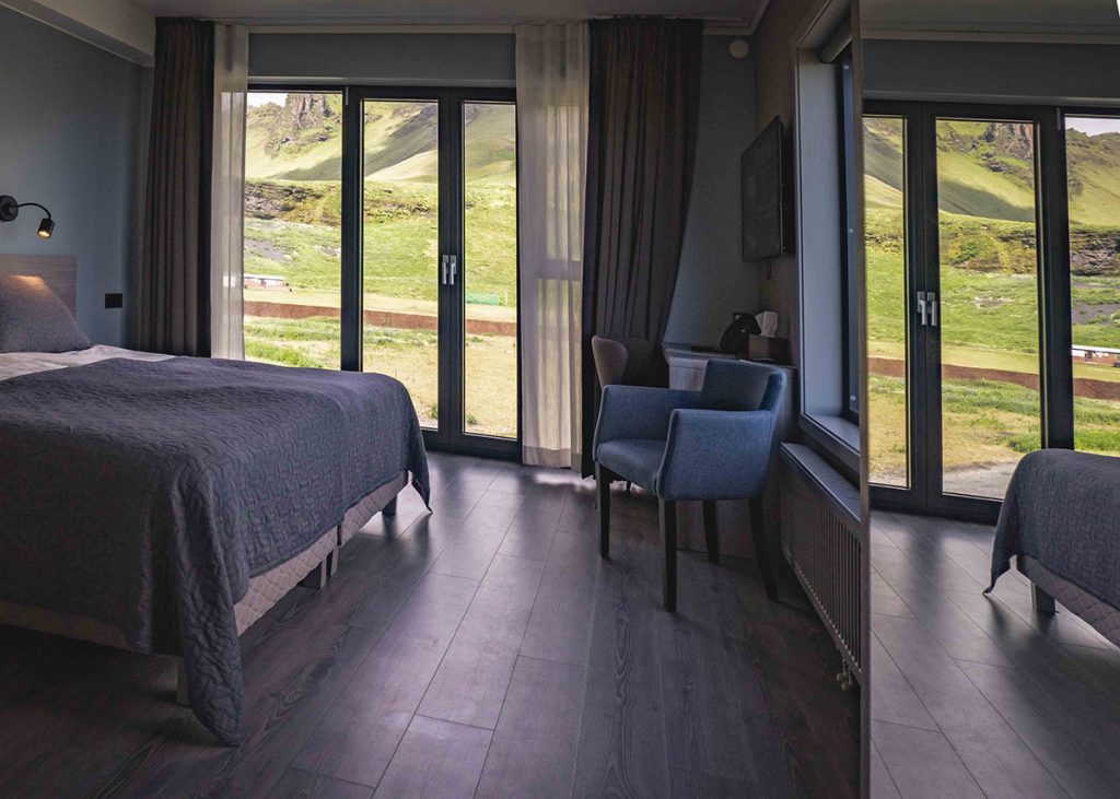A cozy, modern double room at Hotel Kría in Vík, South Iceland, featuring a large bed, minimalist decor, and floor-to-ceiling windows with views of mossy green cliffs and blue skies.