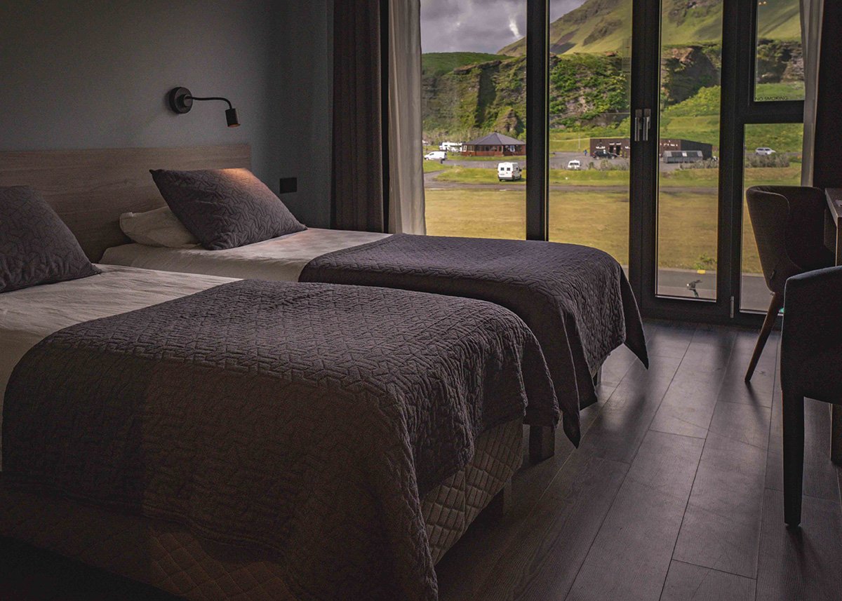 A twin room at Hotel Kría with two neatly made single beds, moody lighting, and expansive views of green mountains, campervans, and small buildings through large glass doors.