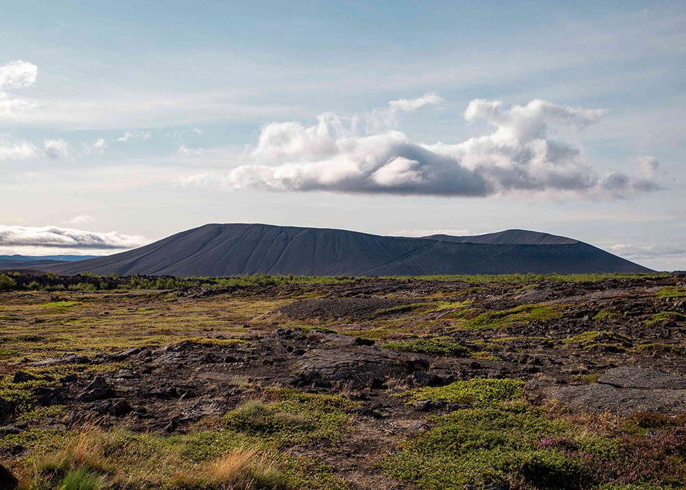Hverfjall, a large volcanic crater in Iceland, viewed from a distance with grassy foreground and a cloudy sky.