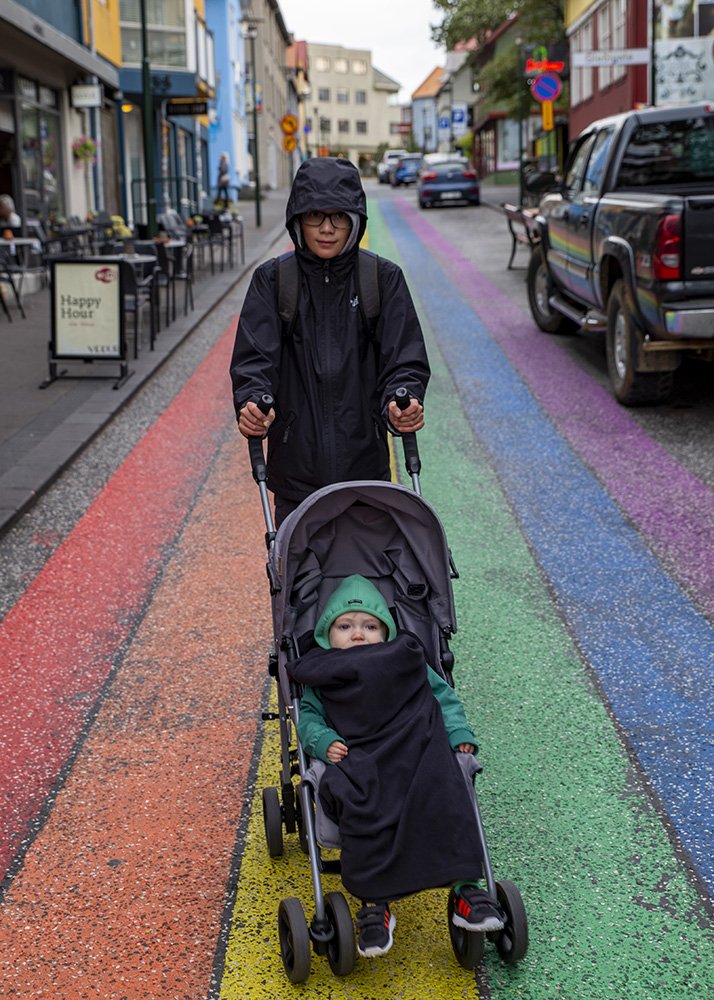 A teenage boy in a black jacket pushing a baby in a stroller down a rainbow-painted street in a colorful urban area.