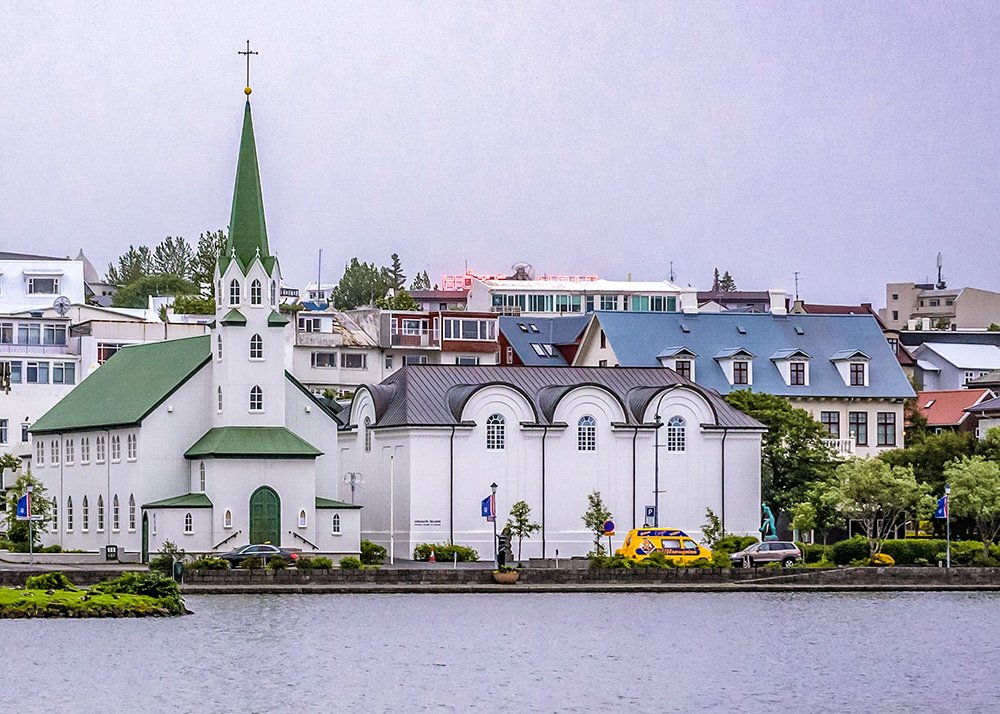 A white church with a green roof and steeple, situated by a lakeside in the cityscape of Reykjavik