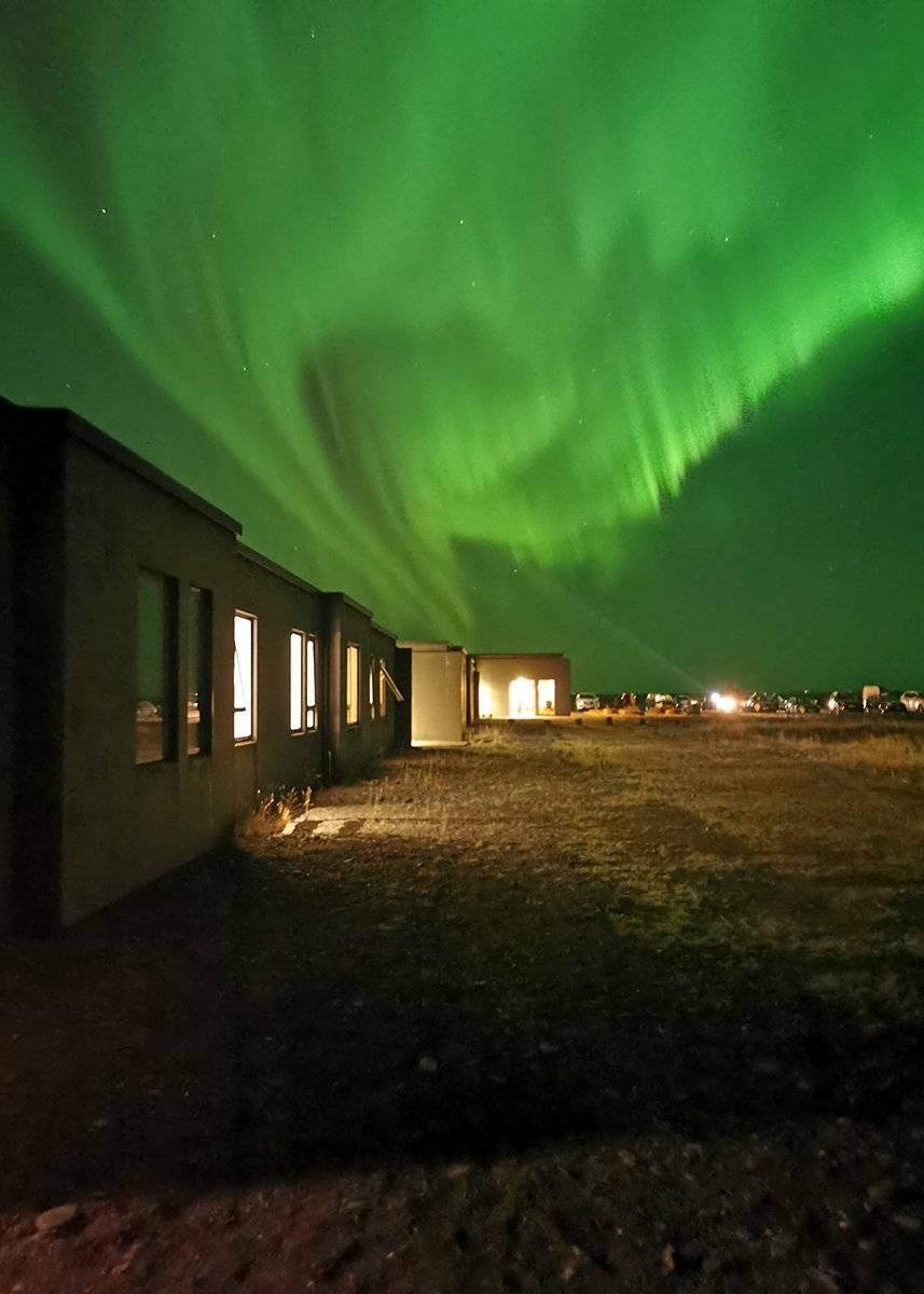 UMI Hotel illuminated at night under a vivid green aurora borealis in the dark sky, with the hotel's windows warmly glowing and parked cars faintly visible in the distance.