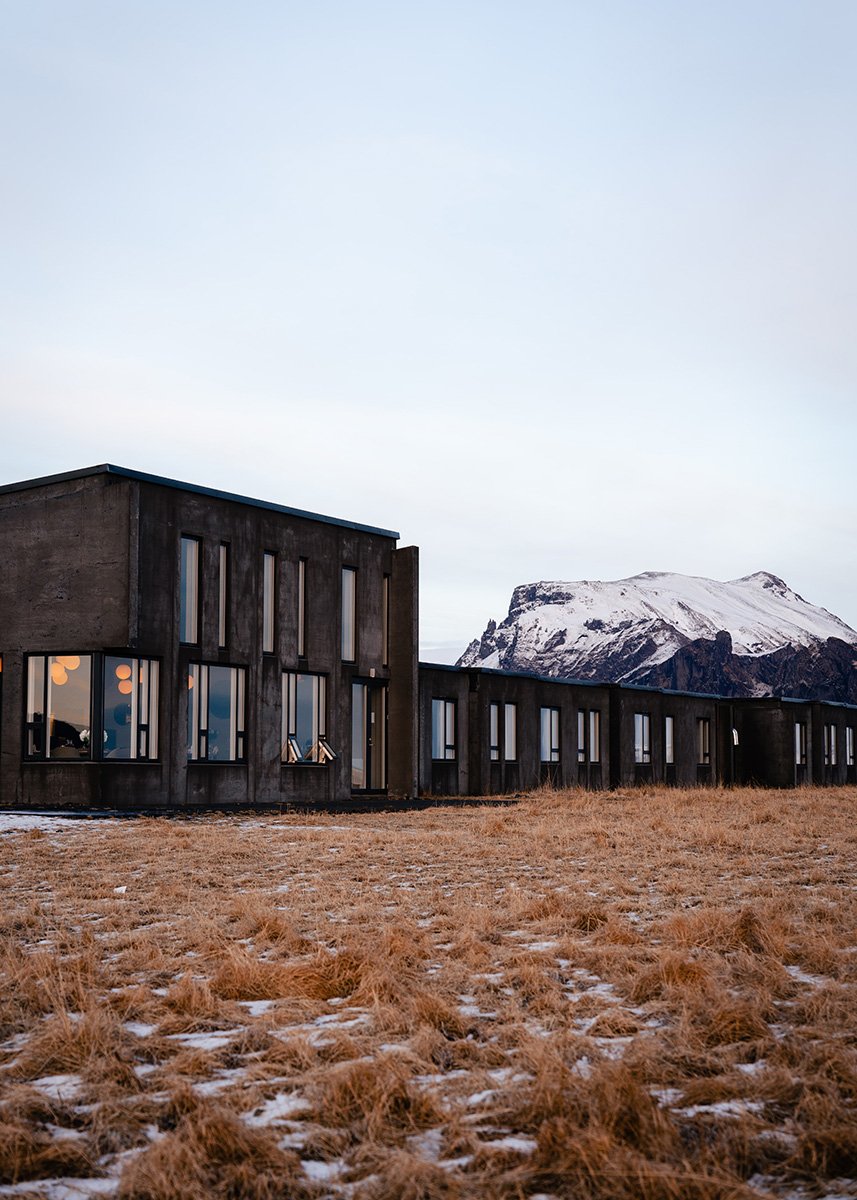 A minimalist concrete building of UMI Hotel in South Iceland, set against a dramatic backdrop of a snow-capped mountain under a pale blue sky, with patchy snow over dry golden grass in the foreground.