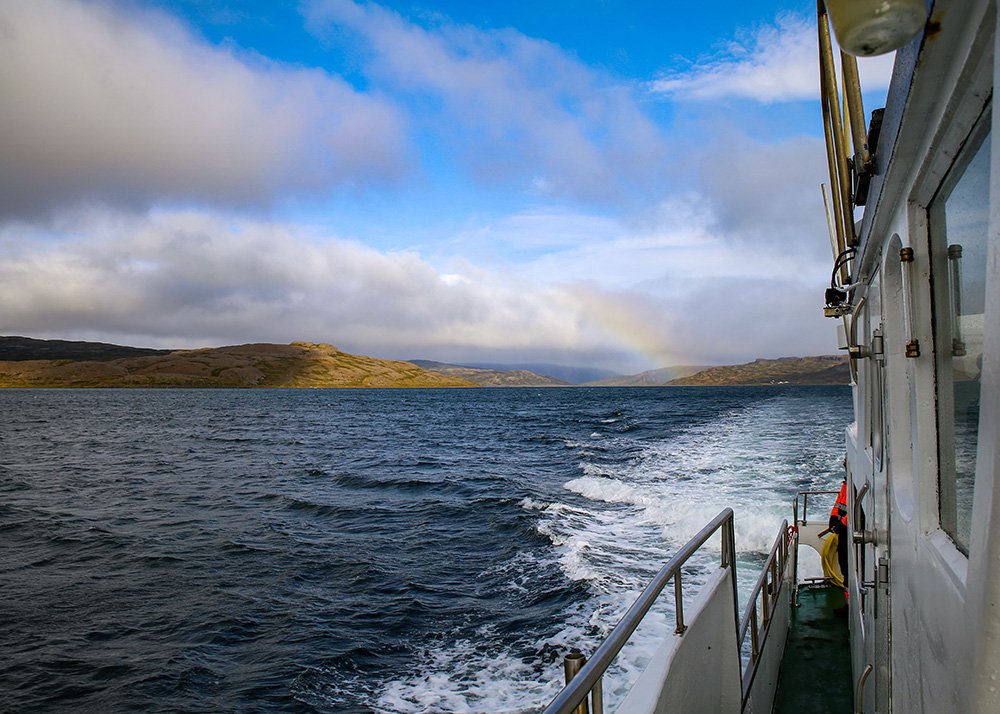 A scenic view from a boat of the ocean and surrounding land with a faint rainbow in the distance under a partly cloudy sky.