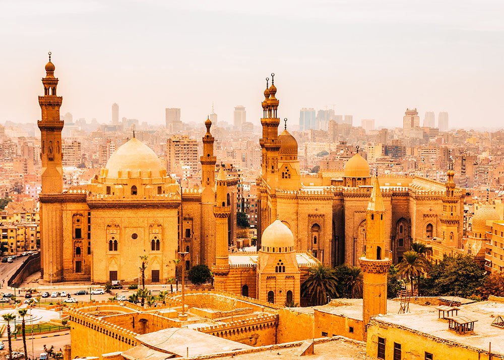 A panoramic view of the Mosque of Sultan Hassan and Al-Rifa'i Mosque in Cairo, with a hazy cityscape in the background.
