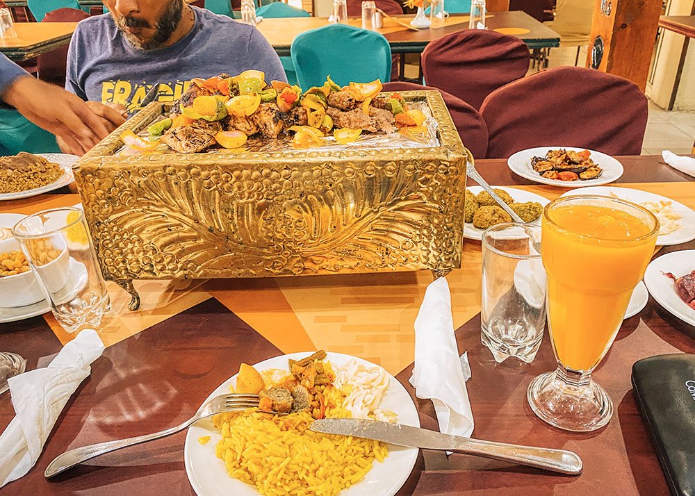 A lavish table setting featuring a large, ornate golden tray filled with assorted food, with plates of food and a glass of orange juice around it.